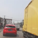 Mobil sedan merah harus mengabil arah keluar jalur kekiri pada mobil truk panjang warna kuning.